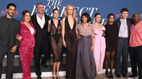 Getty Images The cast of The Perfect Couple, left to right, Ishan Hutter, Donna Lynne Champlin, Liev Schreiber, Megan Fahey, Nicole Kidman, Suzanne Bier, Eva Hewson, Mia Isaacs, Sam Nivola and Michael Beach. At the Los Angeles premiere, with "A perfect pair" written in white behind the actor on a blue background.