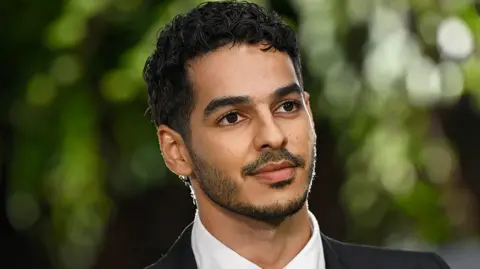 Getty Images Male actor Ishaan Khatter looks off at the premiere wearing a white shirt and black blazer. The green trees in the background are blurry and out of focus.